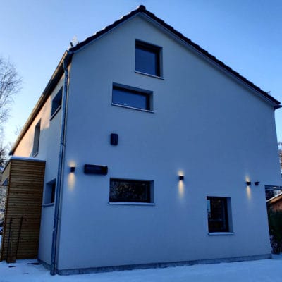 Moderne Hausfassade mit Wandbeleuchtung im Schnee neben einer hohen Hecke im Winter