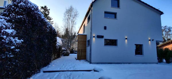 Moderne Hausfassade mit Wandbeleuchtung im Schnee neben einer hohen Hecke im Winter