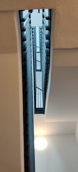 Blick durch das Treppenhaus mit vertikaler Öffnung; SHI Gebäudetechnik installiert runde LED-Wandleuchte und lineare Lichtführung für sichere Ausleuchtung.