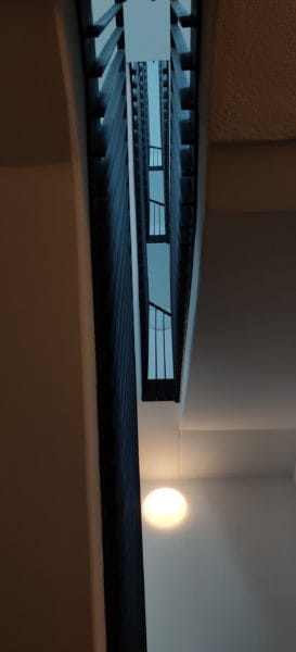 Blick durch das Treppenhaus mit vertikaler Öffnung; SHI Gebäudetechnik installiert runde LED-Wandleuchte und lineare Lichtführung für sichere Ausleuchtung.