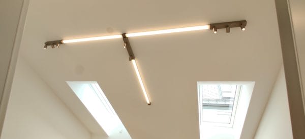 Deckenbeleuchtung aus drei linearen LED-Lichtbändern mit integrierten Spots zwischen zwei Dachfenstern, montiert von SHI Gebäudetechnik.
