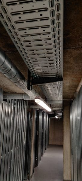Kabeltrasse mit sauber geführten Leitungen und Feuchtraum-LED-Leuchte über Abteilen im Keller, installiert von SHI Gebäudetechnik.