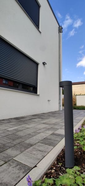 Gepflasterte Terrasse am Einfamilienhaus in Dachau mit grauer Fassade, Rollläden, Wandaußenleuchte und Pollerleuchte im Vordergrund