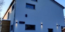 Moderne Hausfassade mit Wandbeleuchtung im Schnee neben einer hohen Hecke im Winter